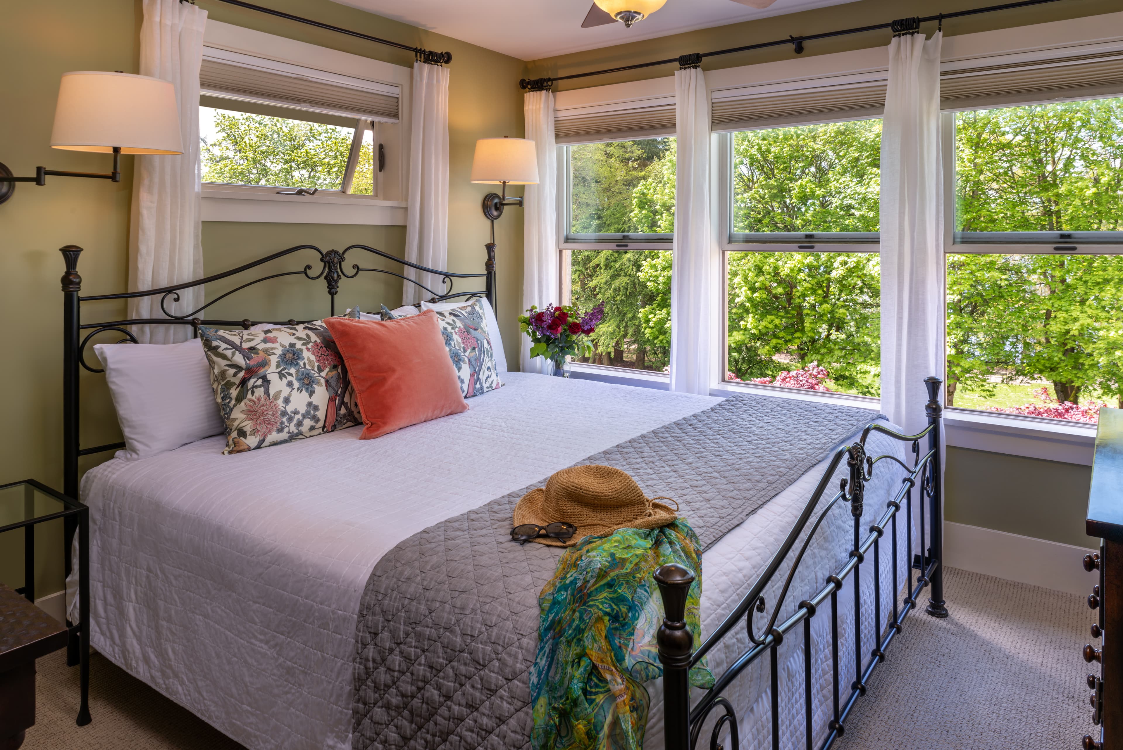 A cozy bedroom with a queen-sized bed, adorned with floral and solid pillows, a quilted gray blanket, and an iron bed frame. Large windows with white curtains offer views of lush greenery, filling the room with natural light.