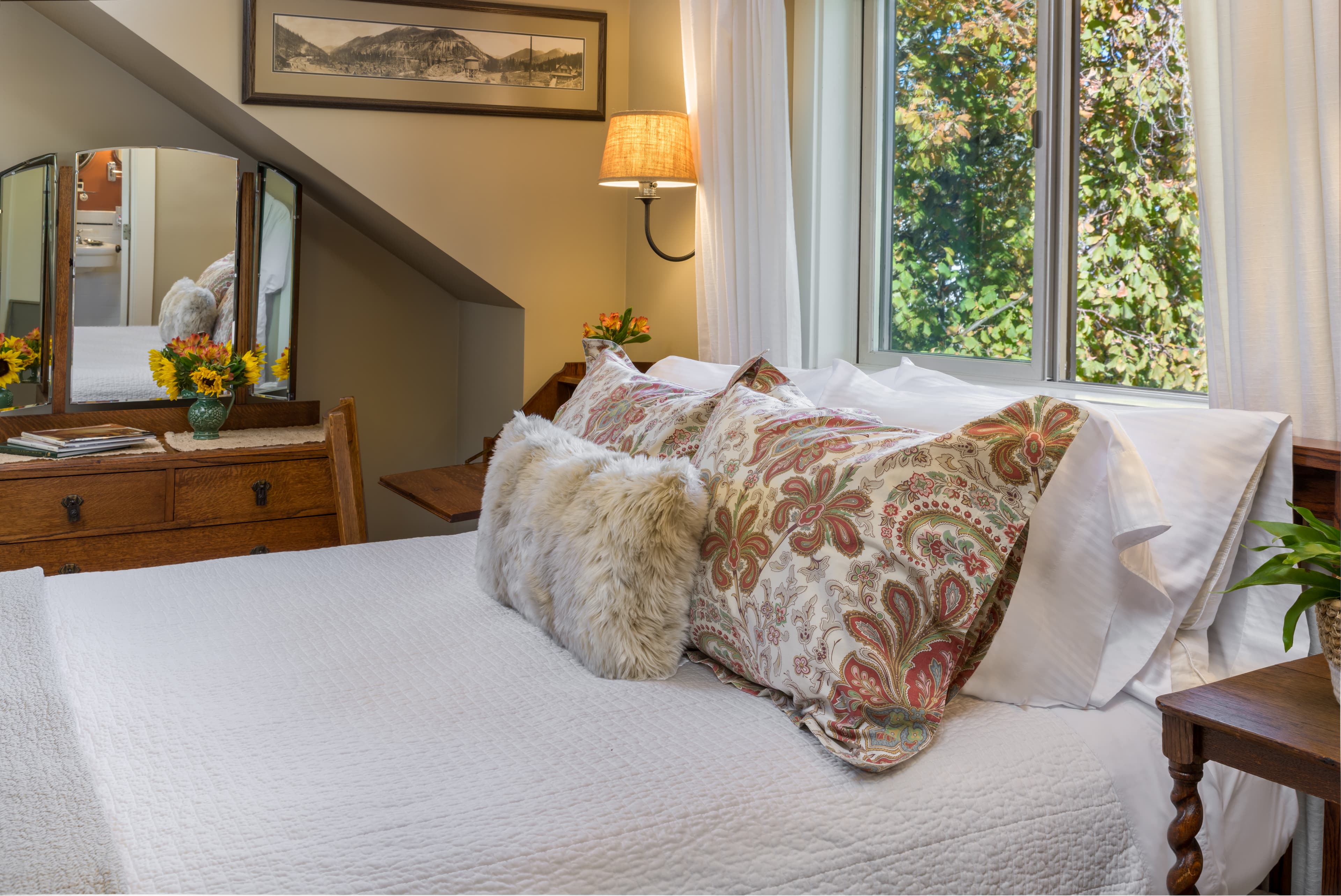 Cozy bedroom with a neatly made bed, patterned pillows, a bedside table, lamp, and a window with a view of trees.