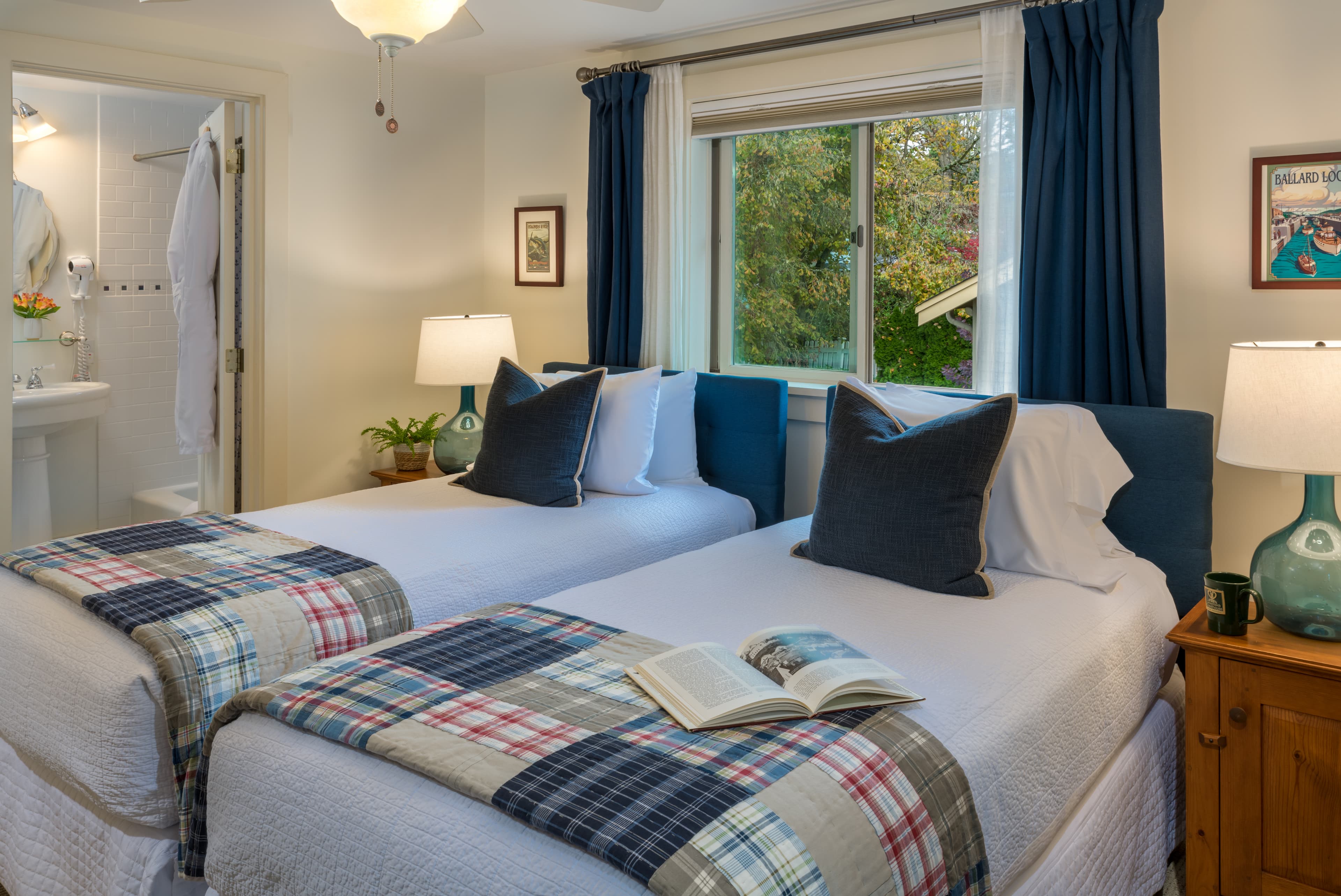 A cozy bedroom with twin beds, plaid covers, blue curtains, open book, and a view of greenery outside.