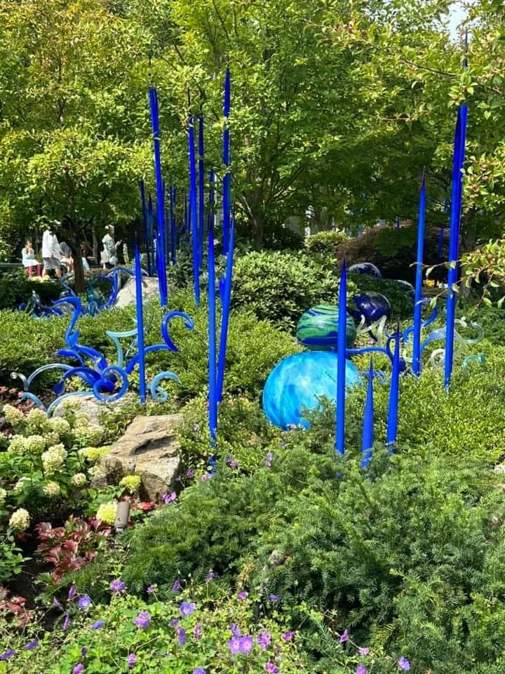 Vibrant blue glass sculptures surrounded by lush greenery and colorful flowers in a garden setting.