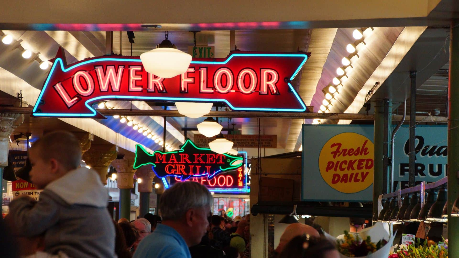 A bustling market interior featuring colorful neon signs, including "Lower Floor" and "Fresh Picked Daily."